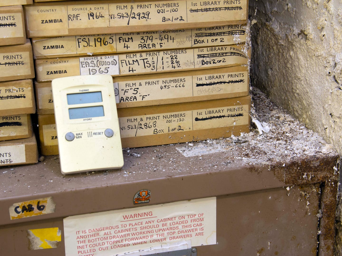 A pile of slender cardboard boxes marked with flight information, stacked on a metal filing cabinet, next to a damp and peeling wall.