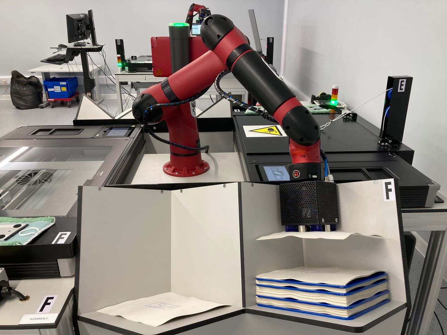 A red robotic arm carefully lifts a single photographic print from a stack of prints towards a large flatbed scanner.