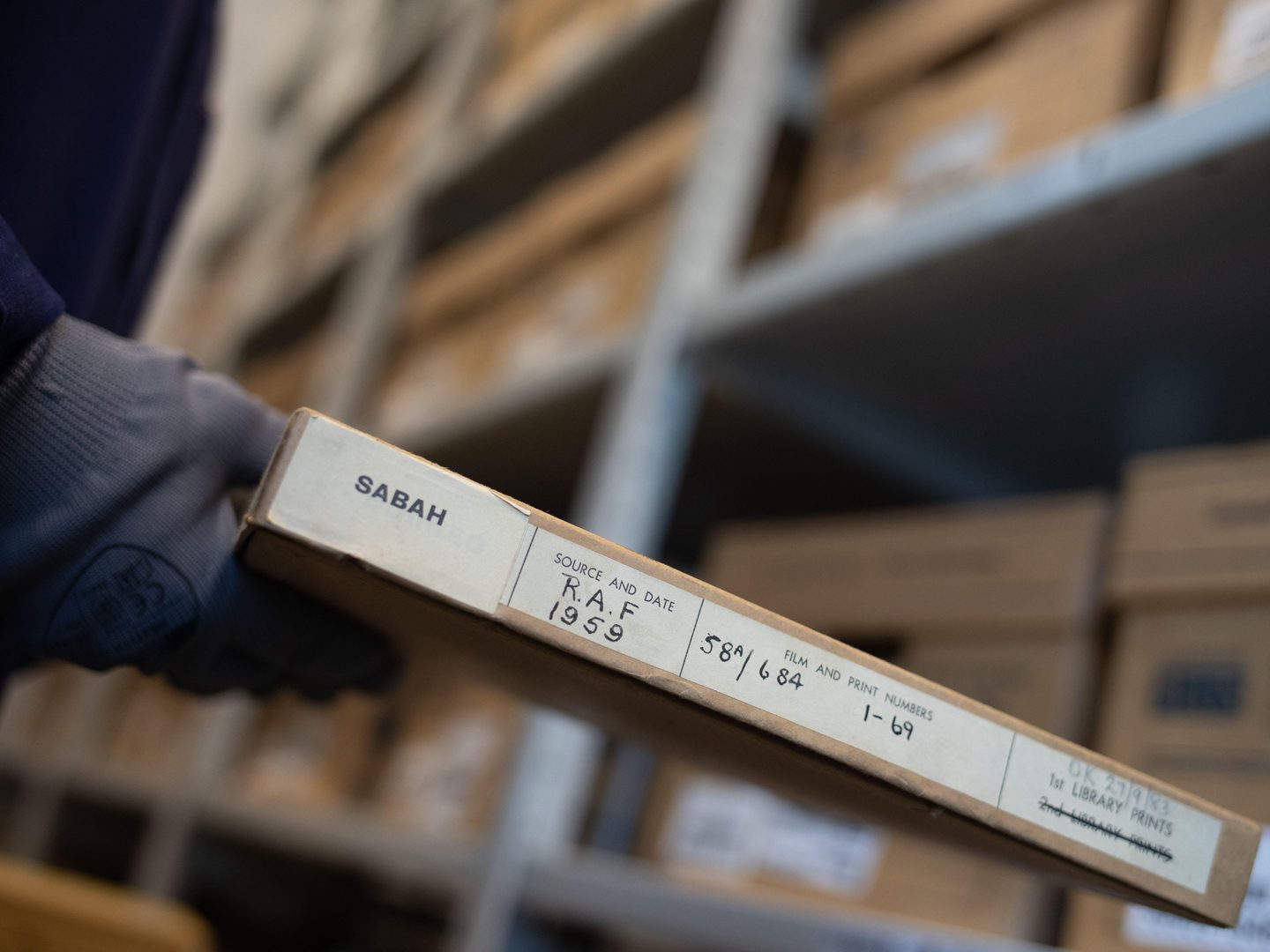 A hand wearing a grey glove holds a slender cardboard box. The box is labelled ‘Sabah’. Other information on the box suggests it contains aerial photos taken by the RAF in 1959. There are more boxes on grey shelves out of focus in the background.
