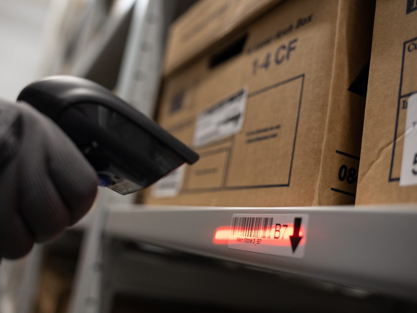 A grey-gloved hand holds a black barcode scanner. The scanner is emitting a red light which has been pointed at a barcode label. The label is attached to a metal shelf holding 2 cardboard boxes.