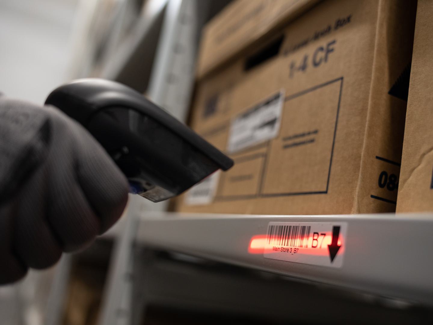 A grey-gloved hand holds a black barcode scanner. The scanner is emitting a red light which has been pointed at a barcode label. The label is attached to a metal shelf holding 2 cardboard boxes.