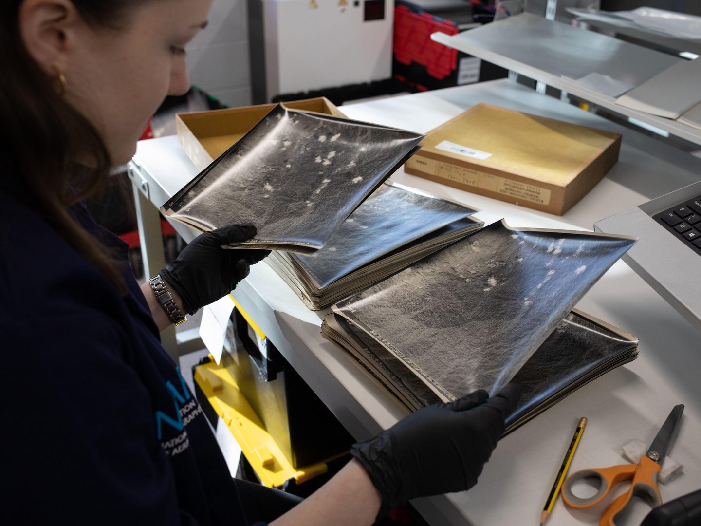 A woman wearing black nitrile gloves looking through a stack of black and white aerial photos. She is standing at a white desk with two stacks of prints in front of her and a print in each hand. The cardboard box for the prints is also visible.