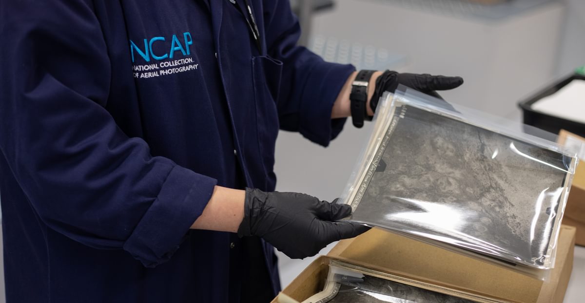 A person wearing a blue lab coat with the NCAP logo holds a black and white aerial photographic print in black-gloved hands.