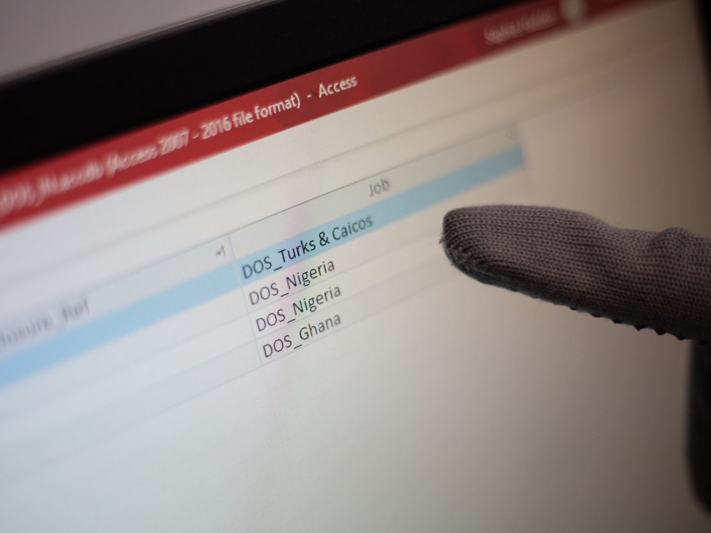 A grey-gloved finger points at a table on a computer monitor. Only one column of the table is visible, in which the entries read DOS Turks & Ciacos, DOS Nigeria and DOS Ghana.