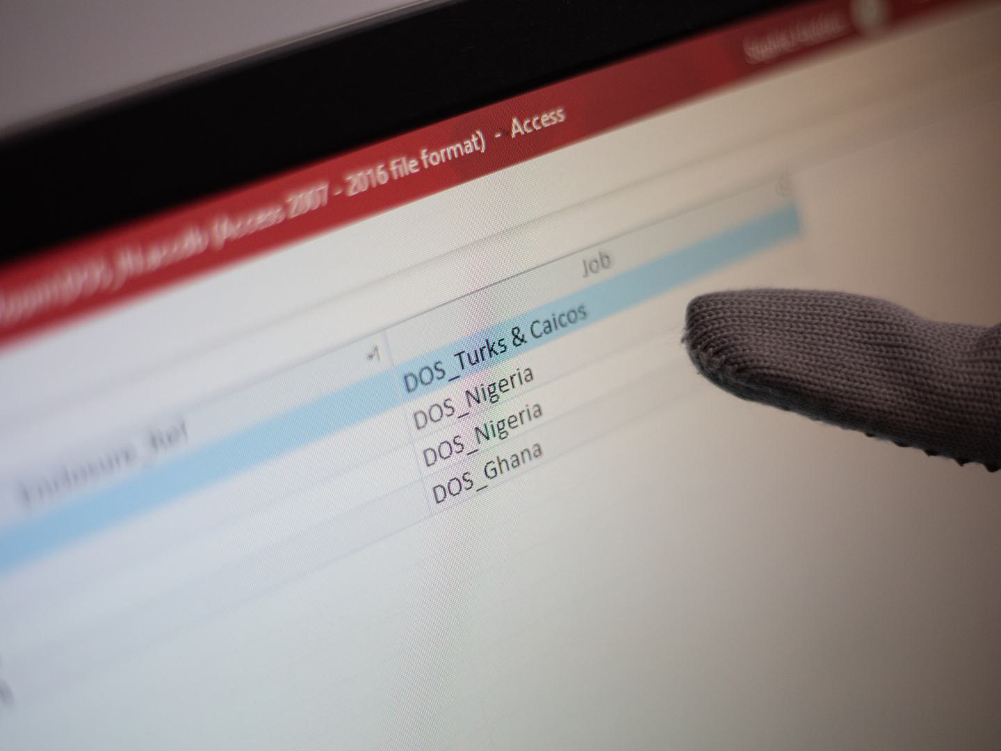 A grey-gloved finger points at a table on a computer monitor. Only one column of the table is visible, in which the entries read DOS Turks & Ciacos, DOS Nigeria and DOS Ghana.
