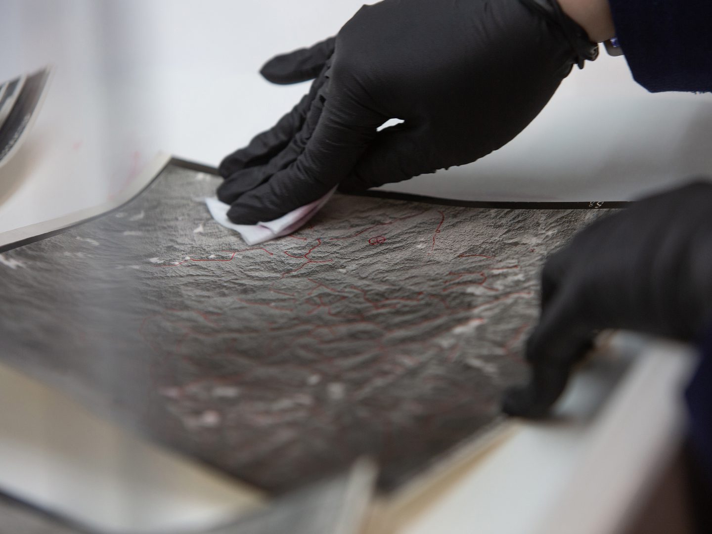 A close-up of a black and white vertical aerial photo of a landscape. Only a small amount of the photo is in focus. A black-gloved hand wipes the photo with a white cloth. A second black-gloved hand holds the photo flat.