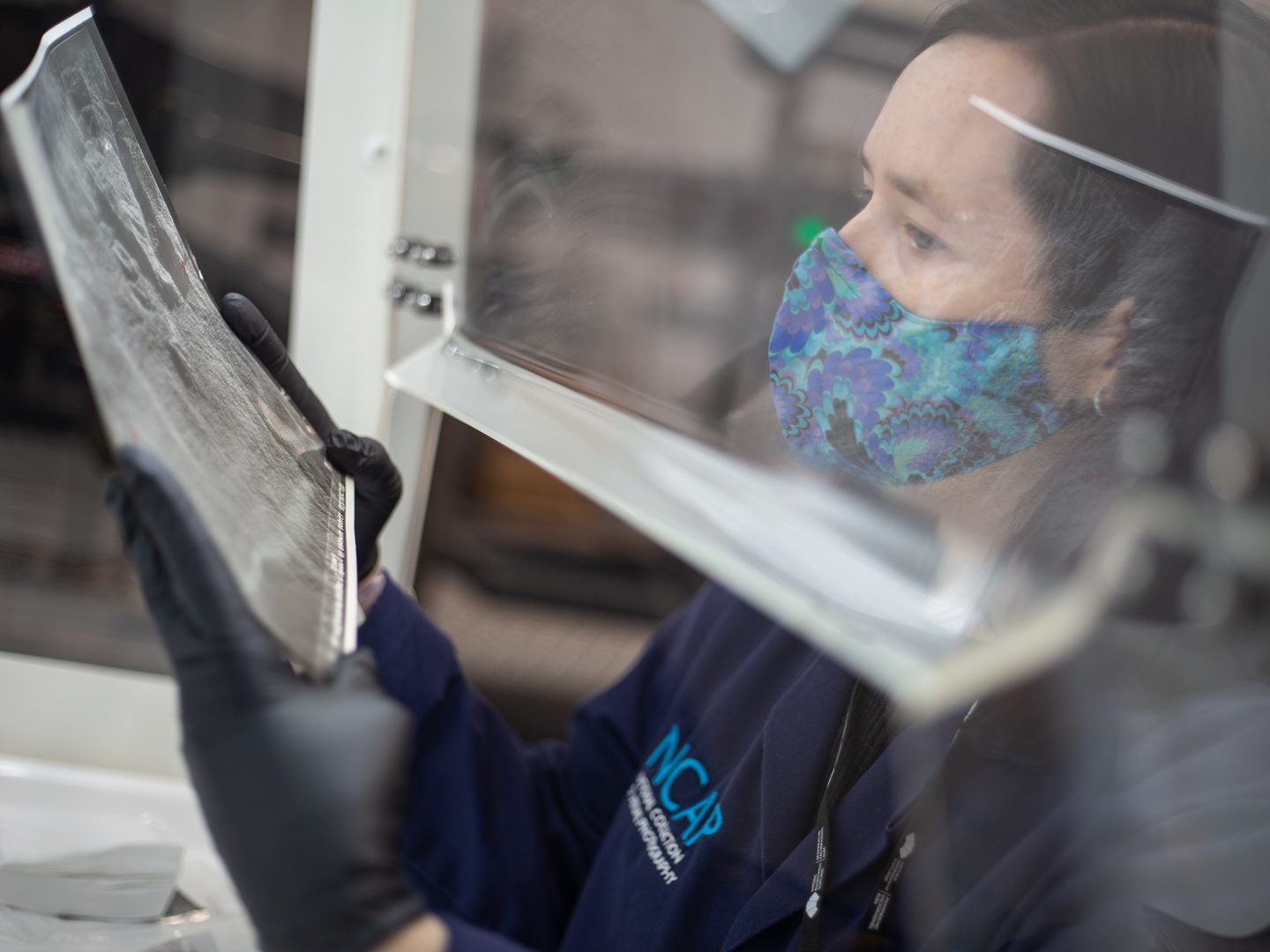 A woman with dark hair and wearing a colourful floral face mask holds up a black and white aerial photo inside a fume cabinet. She is separated from the photo by the Perspex side of the fume cabinet. She is wearing black gloves and a blue lab coat.