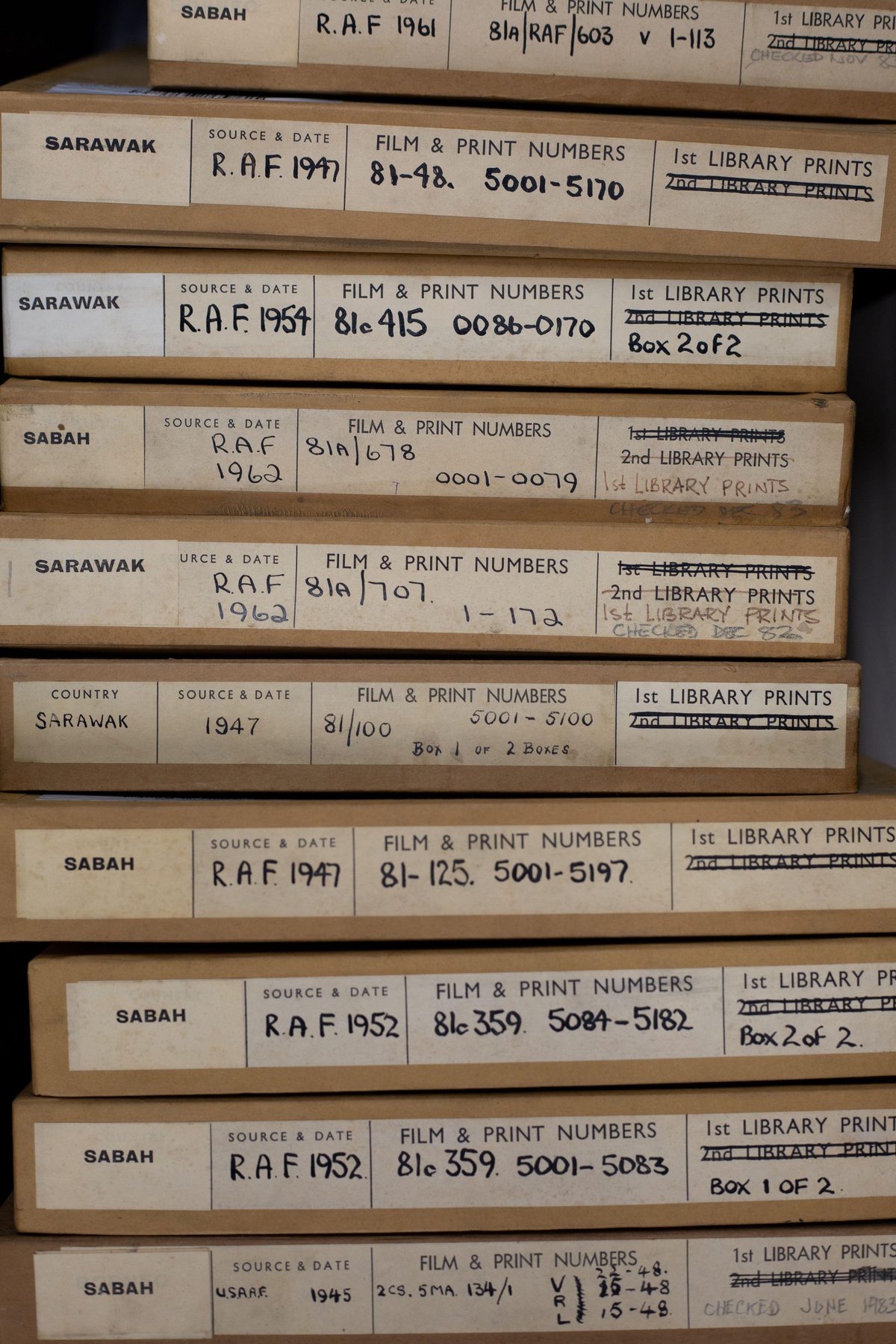 A stack of 10 thin cardboard boxes for aerial photos. The boxes have labels on them indicating the photos were taken in Sarawak and Sabah between 1945 and 1962. The labels contain information about the sorties and the squadrons that took the photos.