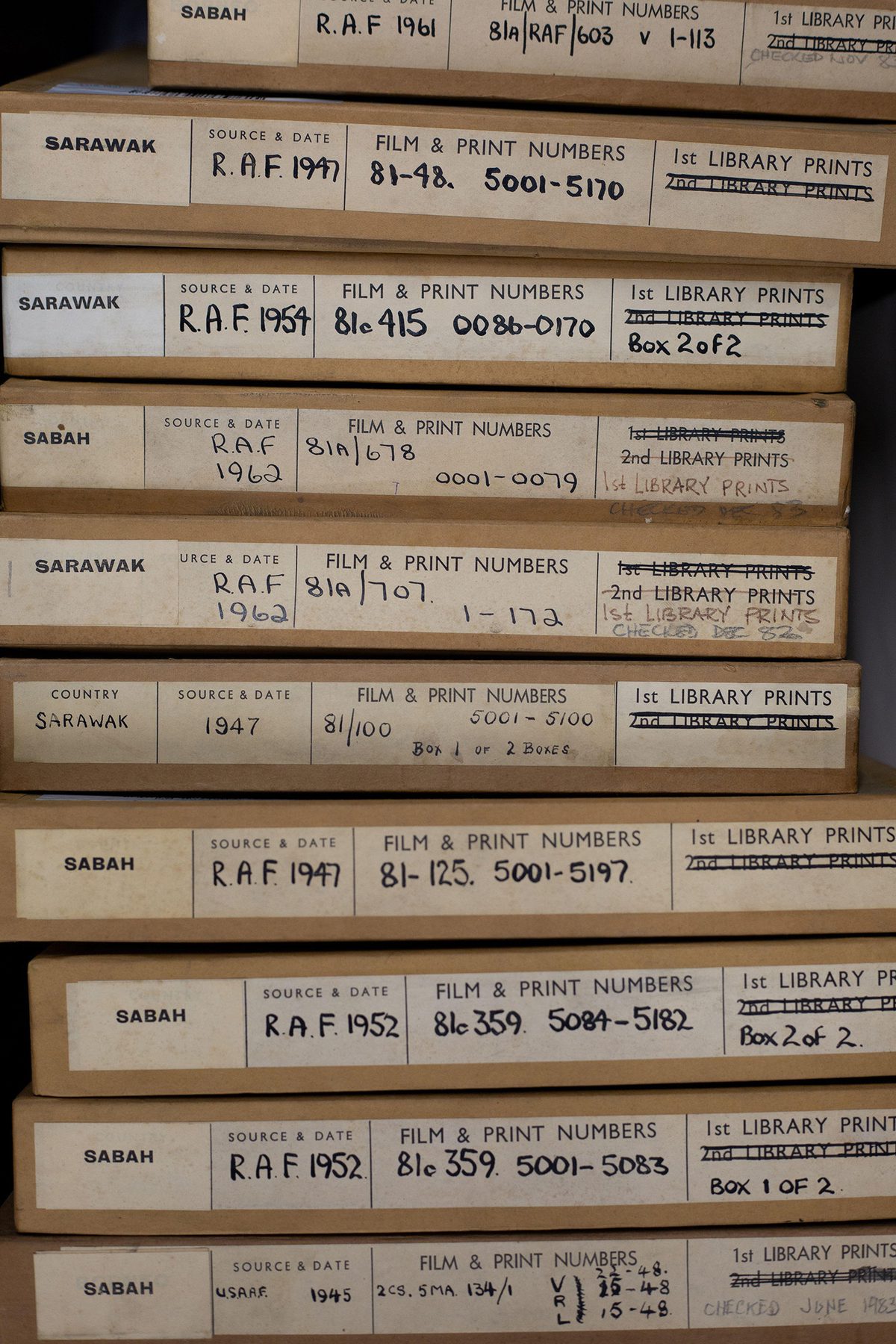 A stack of 10 thin cardboard boxes for aerial photos. The boxes have labels on them indicating the photos were taken in Sarawak and Sabah between 1945 and 1962. The labels contain information about the sorties and the squadrons that took the photos.