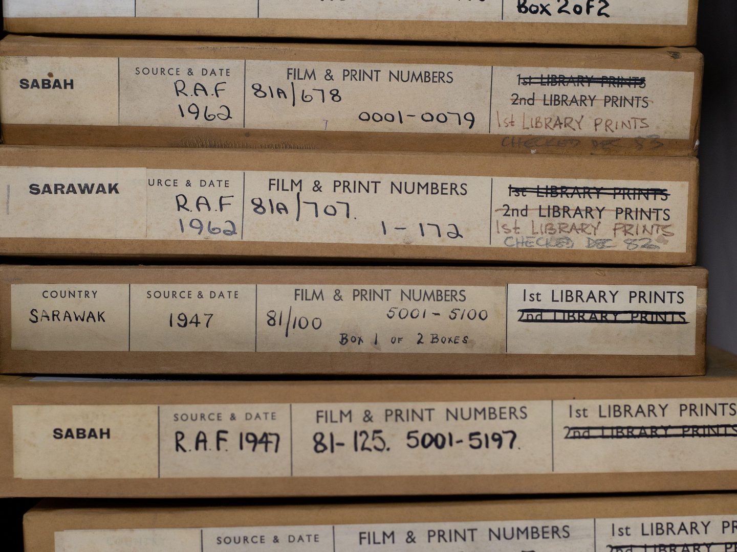 A stack of 10 thin cardboard boxes for aerial photos. The boxes have labels on them indicating the photos were taken in Sarawak and Sabah between 1945 and 1962. The labels contain information about the sorties and the squadrons that took the photos.