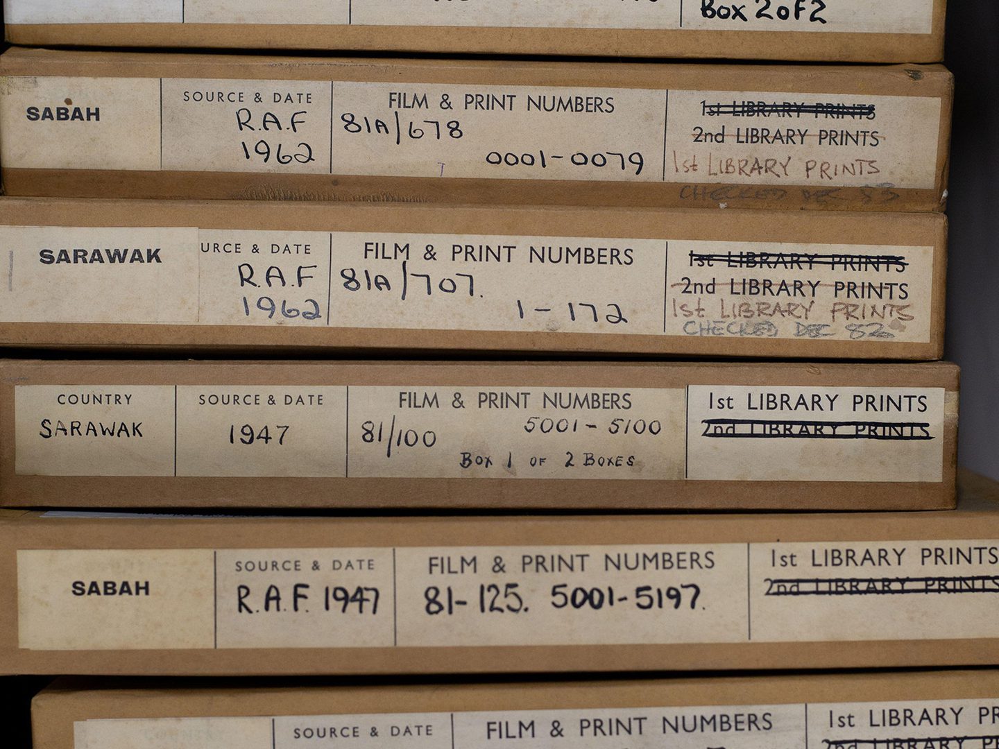 A stack of 10 thin cardboard boxes for aerial photos. The boxes have labels on them indicating the photos were taken in Sarawak and Sabah between 1945 and 1962. The labels contain information about the sorties and the squadrons that took the photos.