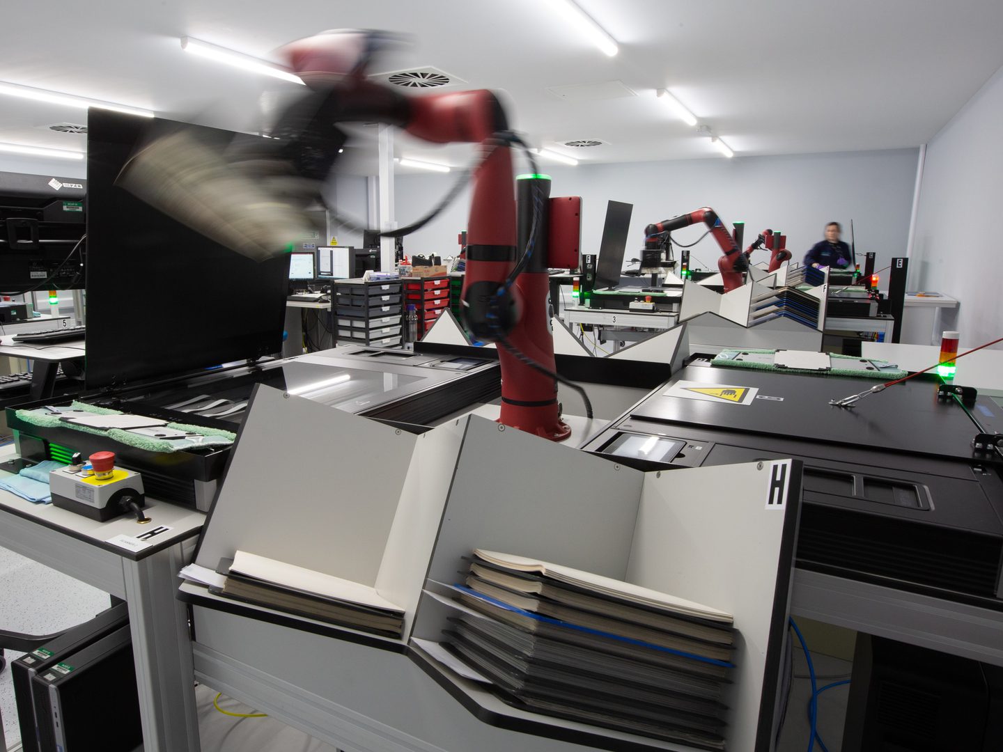 A red robotic arm lifts a photographic print across the centre of the image. The arm sits between two black scanners. Other arms and scanners are visible in the background, along with a person in a blue lab coat facing the camera.