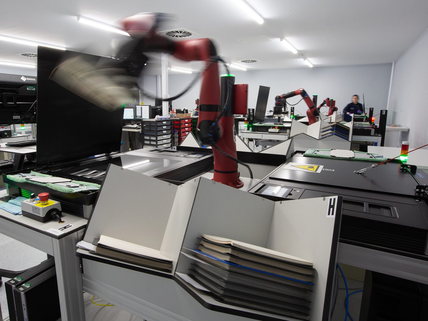 A red robotic arm lifts a photographic print across the centre of the image. The arm sits between two black scanners. Other arms and scanners are visible in the background, along with a person in a blue lab coat facing the camera.