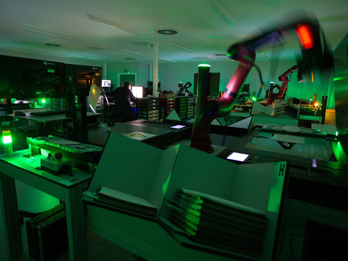 A dark room lit with green lights. In the foreground are two stacks of photographic prints. Above them, a red robotic arm is visible, blurred with movement. In the background, a person with their back to us looks at a bright white screen.