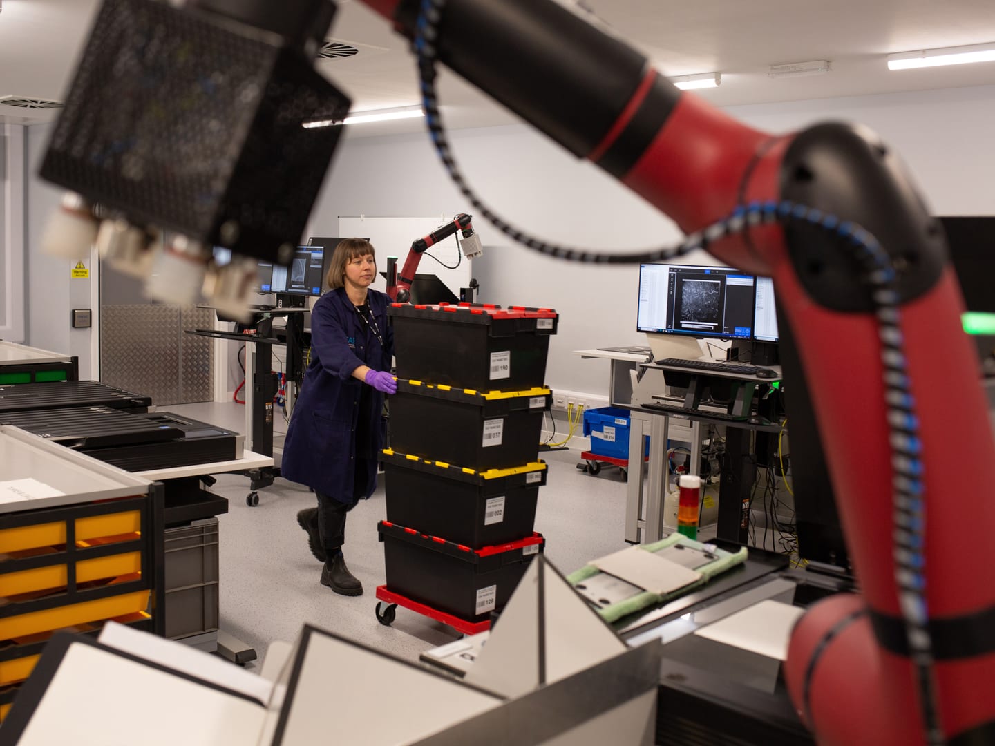 A member of NCAP staff in a blue lab coat pushes a pile of 4 crates stacked on a low red trolley. An out-of-focus red robotic arm frames the foreground.