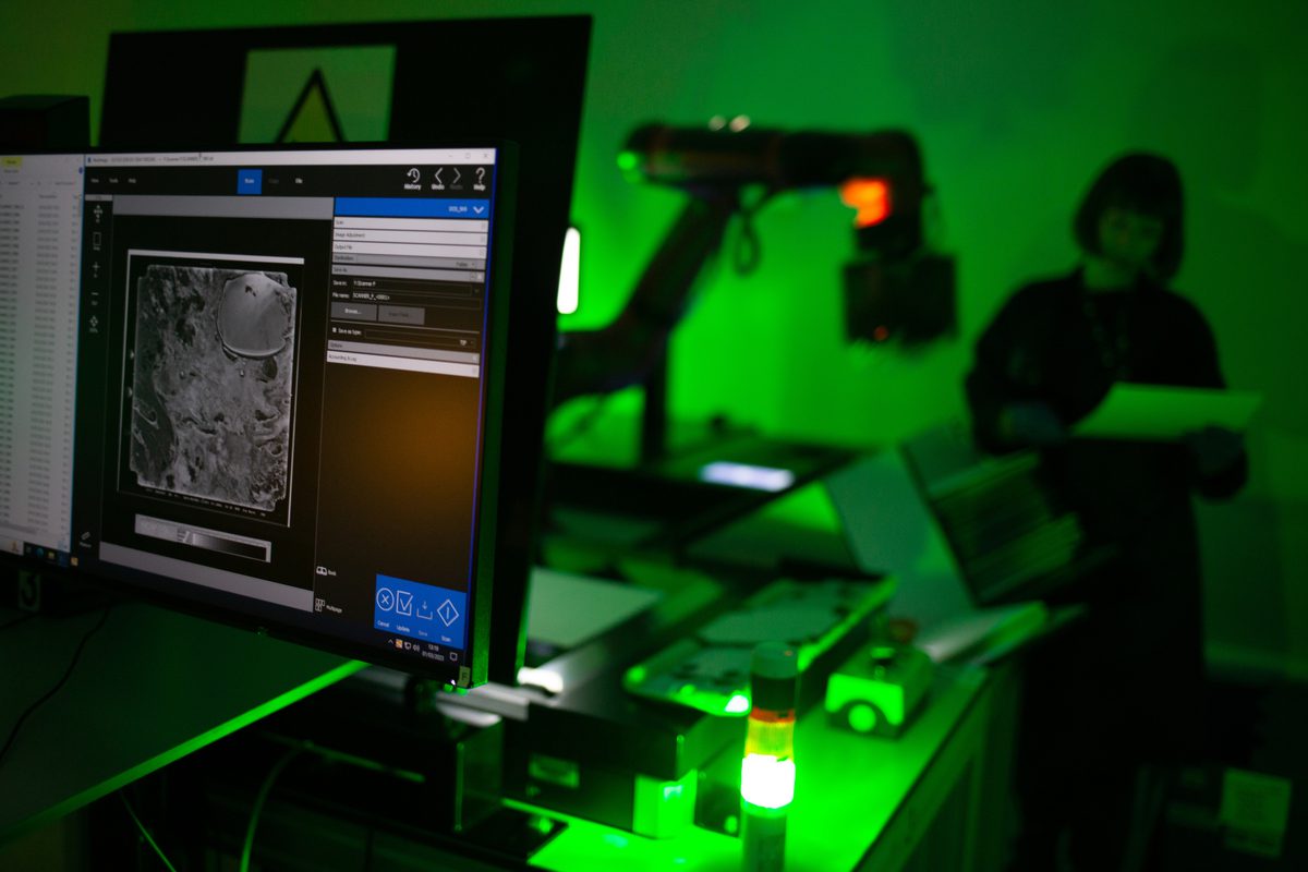 In the foreground is a computer monitor showing an image of a black and white aerial photo. In the background an out-of-focus figure and robotic arm are lit by a green light.