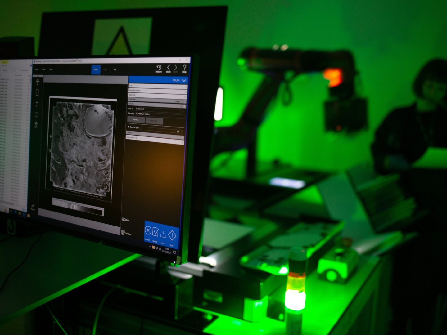 In the foreground is a computer monitor showing an image of a black and white aerial photo. In the background an out-of-focus figure and robotic arm are lit by a green light.