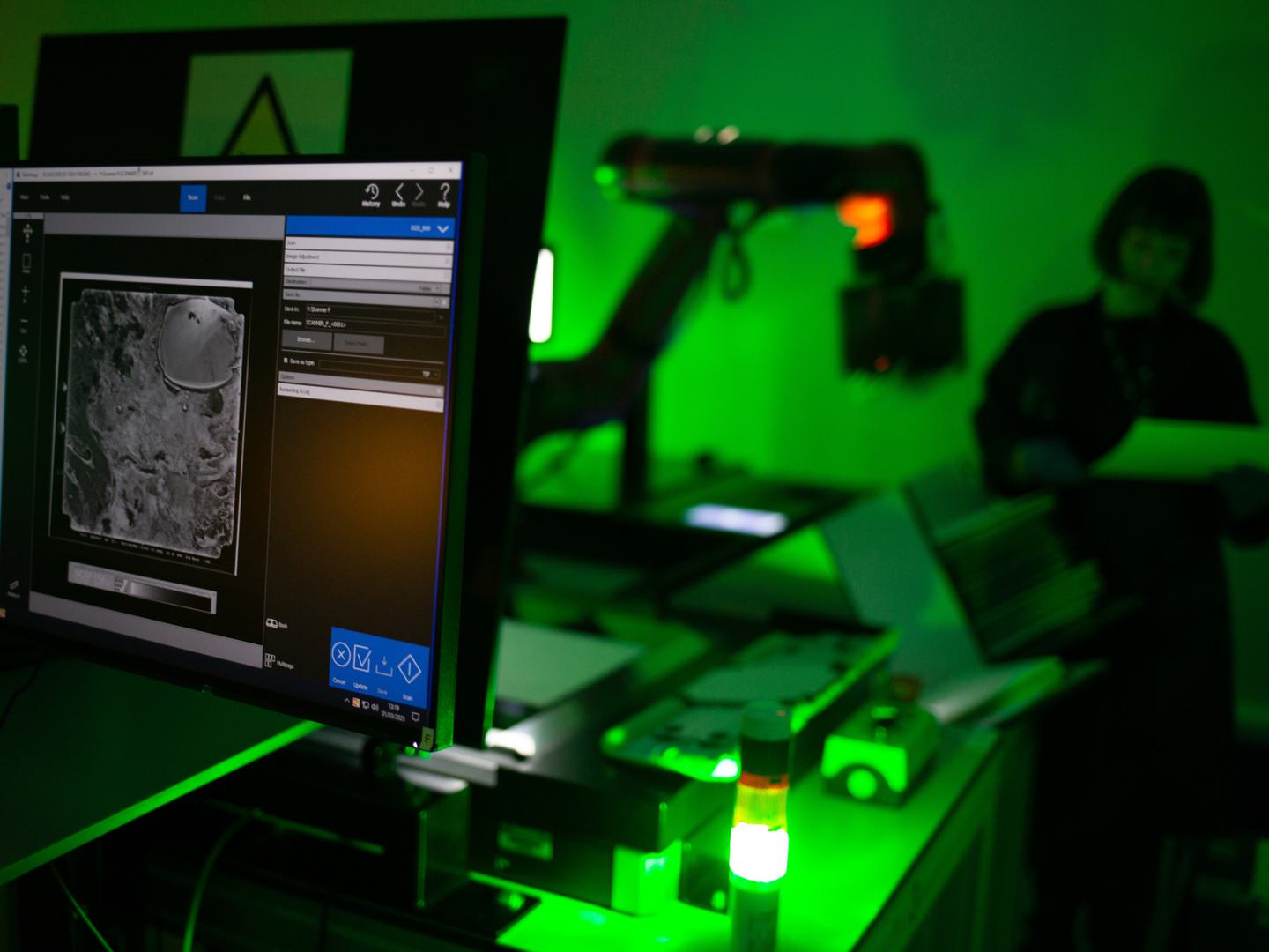 In the foreground is a computer monitor showing an image of a black and white aerial photo. In the background an out-of-focus figure and robotic arm are lit by a green light.