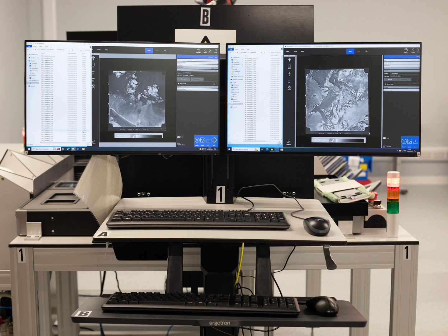 2 computer screens side-by-side. Each shows a different black and white vertical aerial photo of a landscape. 2 keyboards and 2 computer mice sit on the desk in front of the screens.