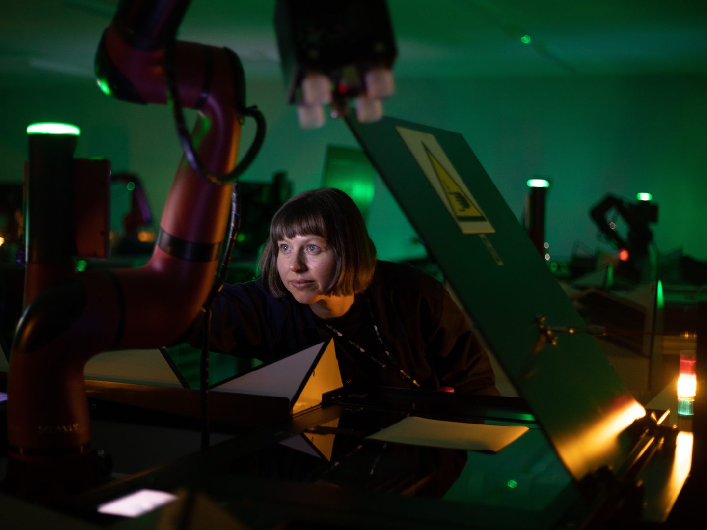A woman with brown hair in a bob leans towards the camera. She is framed by a red robotic arm to the left and the black lid of a scanner to the right. The room is dark and suffused with a greenish glow.