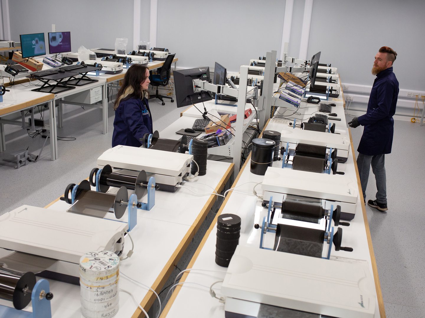 2 rows of flatbed scanners run away from the viewer along white desks. The scanners have rolls of aerial film running through them, suspended on winders either side. 2 people wearing blue lab coats look at computer screens above the scanners.