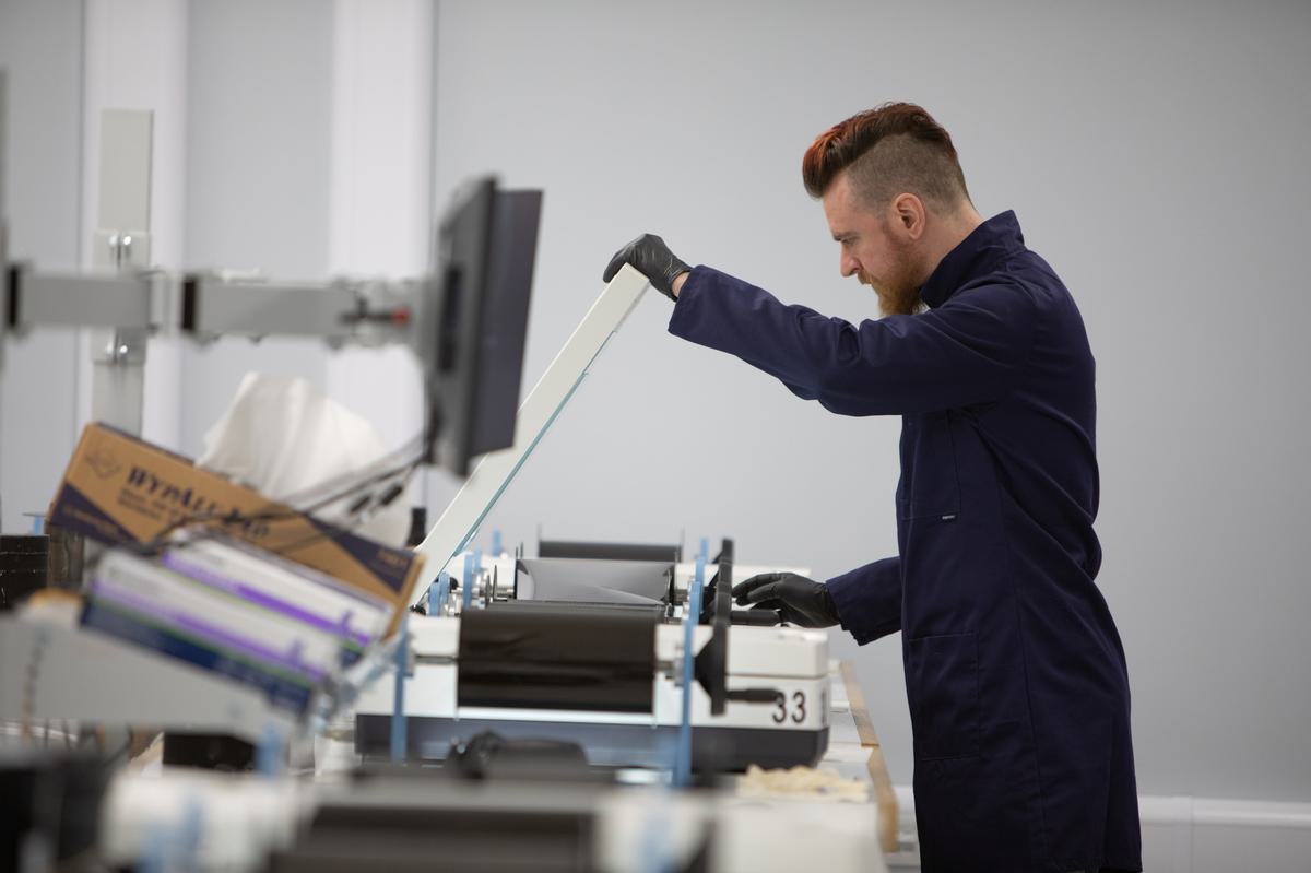 A man with a beard, wearing black gloves and a blue lab coat, lifts the lid of a white flatbed scanner with one hand. With the other hand he winds an aerial photographic film across the scanner bed. An out-of-focus display screen is in the foreground.