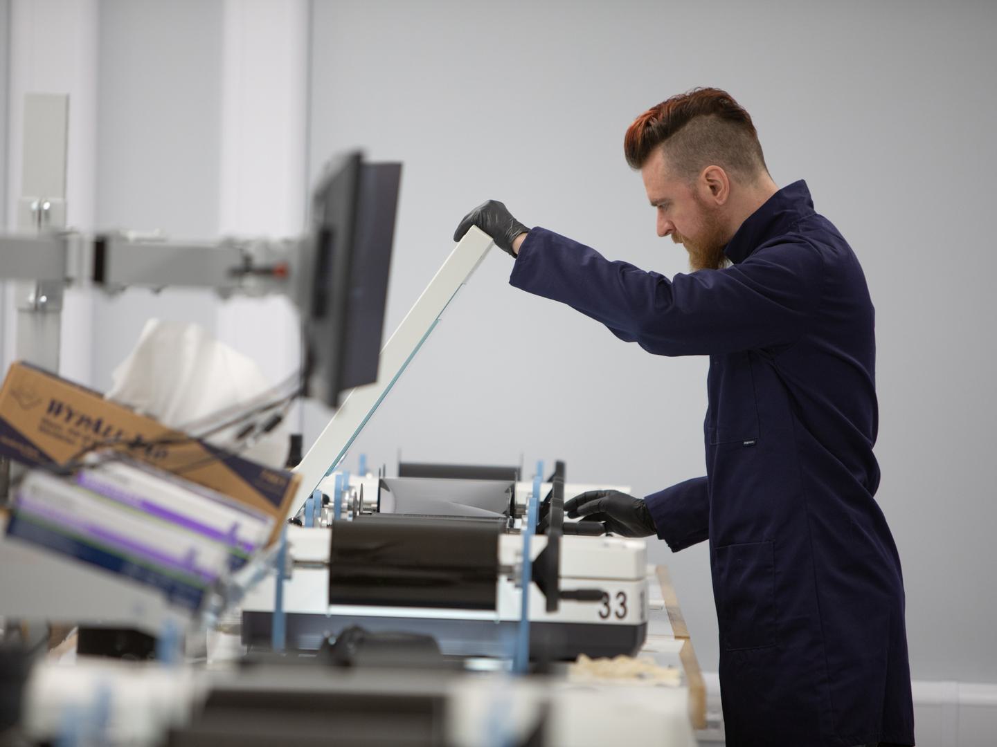 A man with a beard, wearing black gloves and a blue lab coat, lifts the lid of a white flatbed scanner with one hand. With the other hand he winds an aerial photographic film across the scanner bed. An out-of-focus display screen is in the foreground.