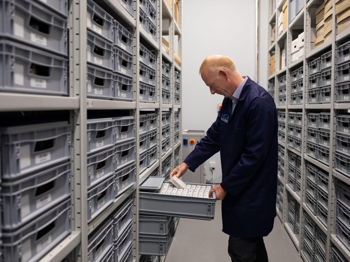 An aisle between two high racks of metal shelving. The shelving is packed with grey plastic trays. A man with red hair in a blue lab coat has pulled one of the trays half off the shelving. He has extracted a small, clear plastic box from the tray.