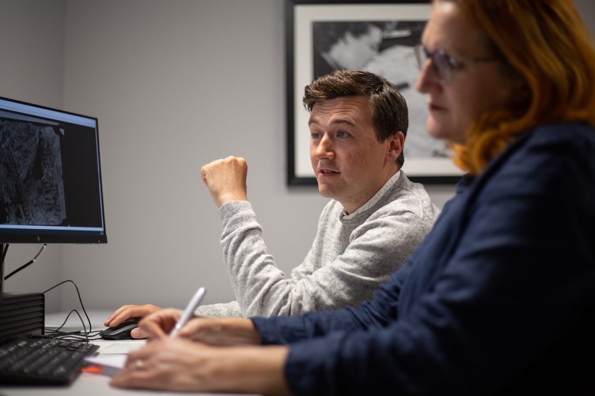 Two people sit at a desk in front of a screen. The screen has a historic black and white aerial photo on it. Out of focus in the foreground is a woman with red hair writing with a pen. In focus in the background is a man in a light grey jumper.