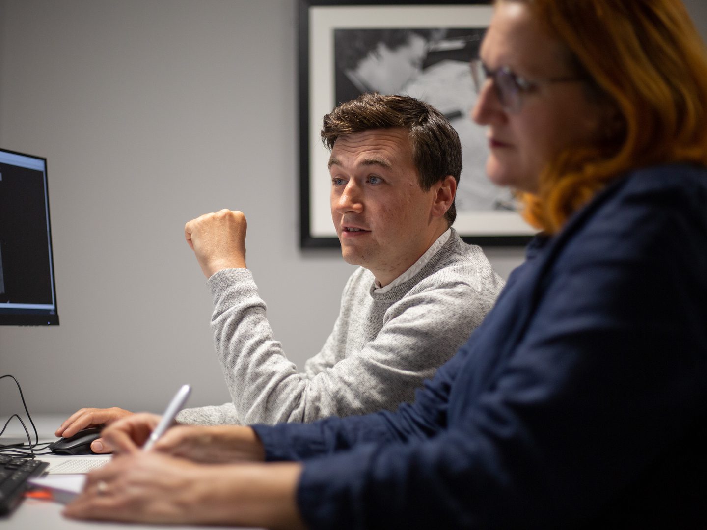 Two people sit at a desk in front of a screen. The screen has a historic black and white aerial photo on it. Out of focus in the foreground is a woman with red hair writing with a pen. In focus in the background is a man in a light grey jumper.