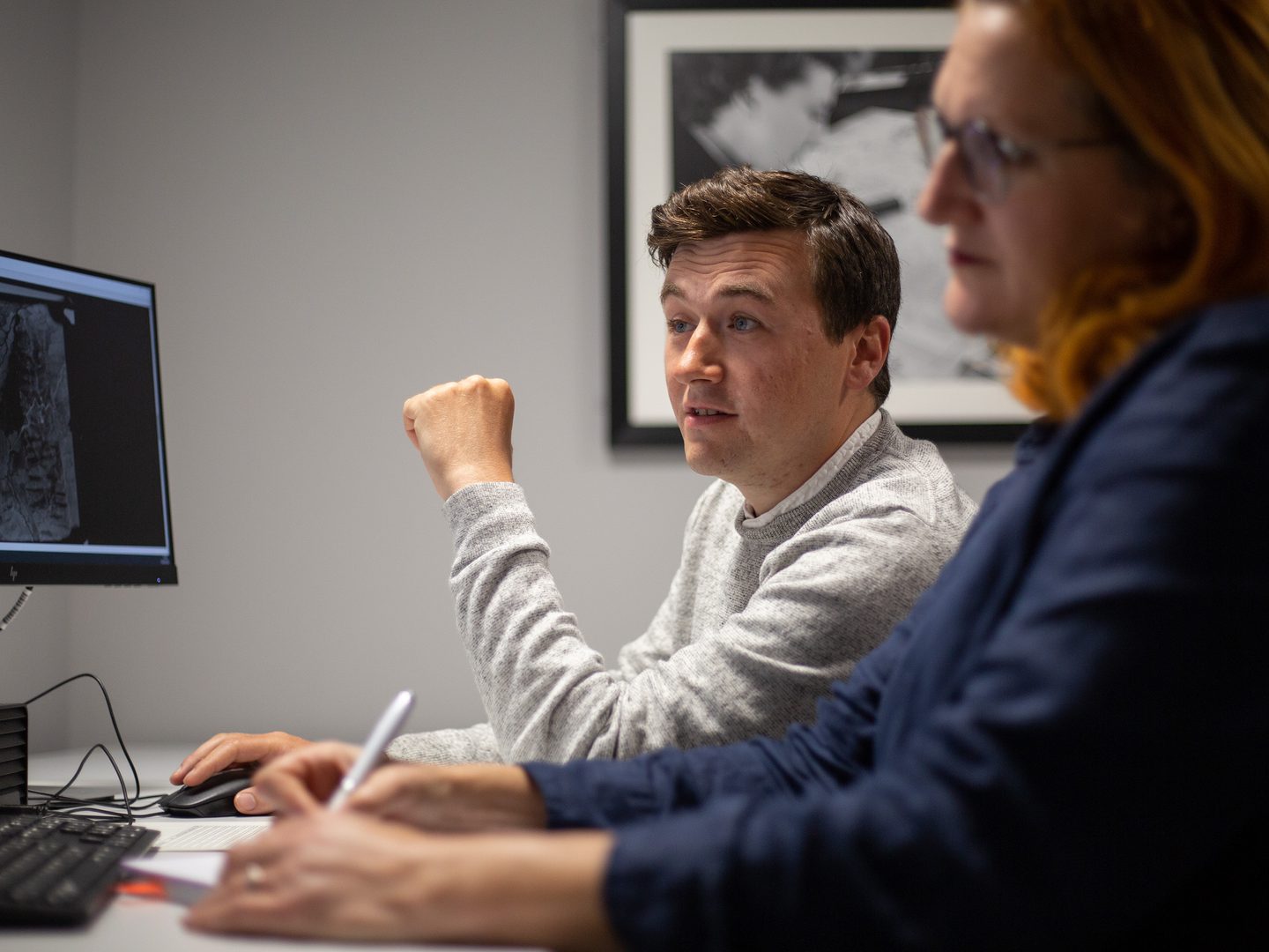 Two people sit at a desk in front of a screen. The screen has a historic black and white aerial photo on it. Out of focus in the foreground is a woman with red hair writing with a pen. In focus in the background is a man in a light grey jumper.
