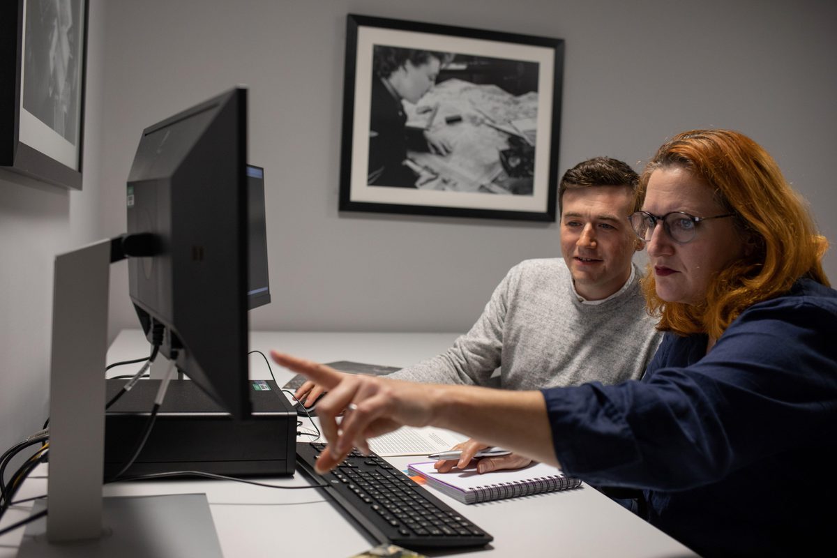 A man and a woman sitting at a desk. The man is wearing a grey jumper. The woman has red hair and is wearing a blue shirt. There is a computer, keyboard and two screens on the desk. The woman is showing the man something on one of the screens.