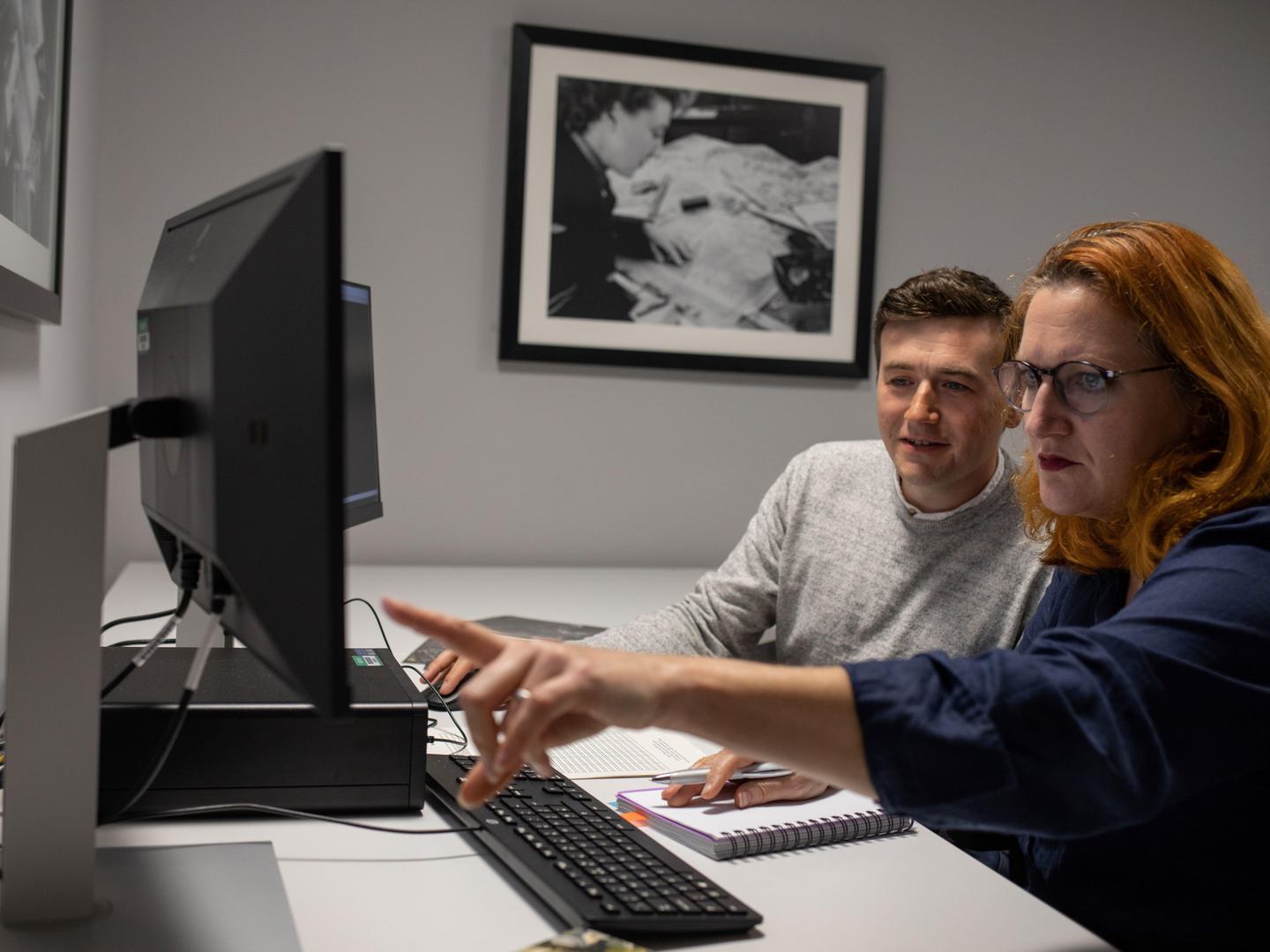 A man and a woman sitting at a desk. The man is wearing a grey jumper. The woman has red hair and is wearing a blue shirt. There is a computer, keyboard and two screens on the desk. The woman is showing the man something on one of the screens.