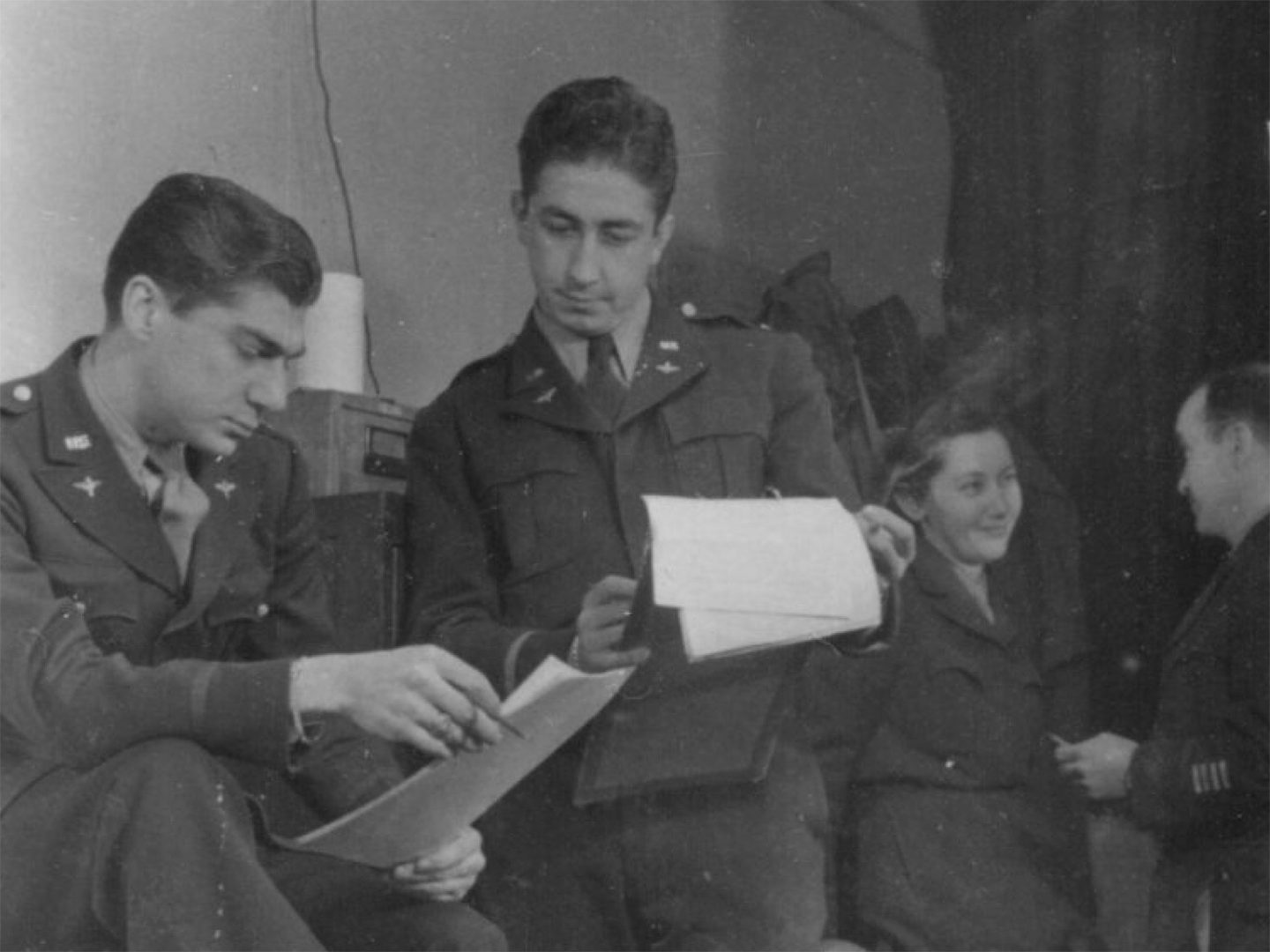 A historic black and white photo of two men with dark hair in US Air Force uniforms consulting some paperwork. One man holds a smoking cigarette. In the background a third man and a woman are talking.