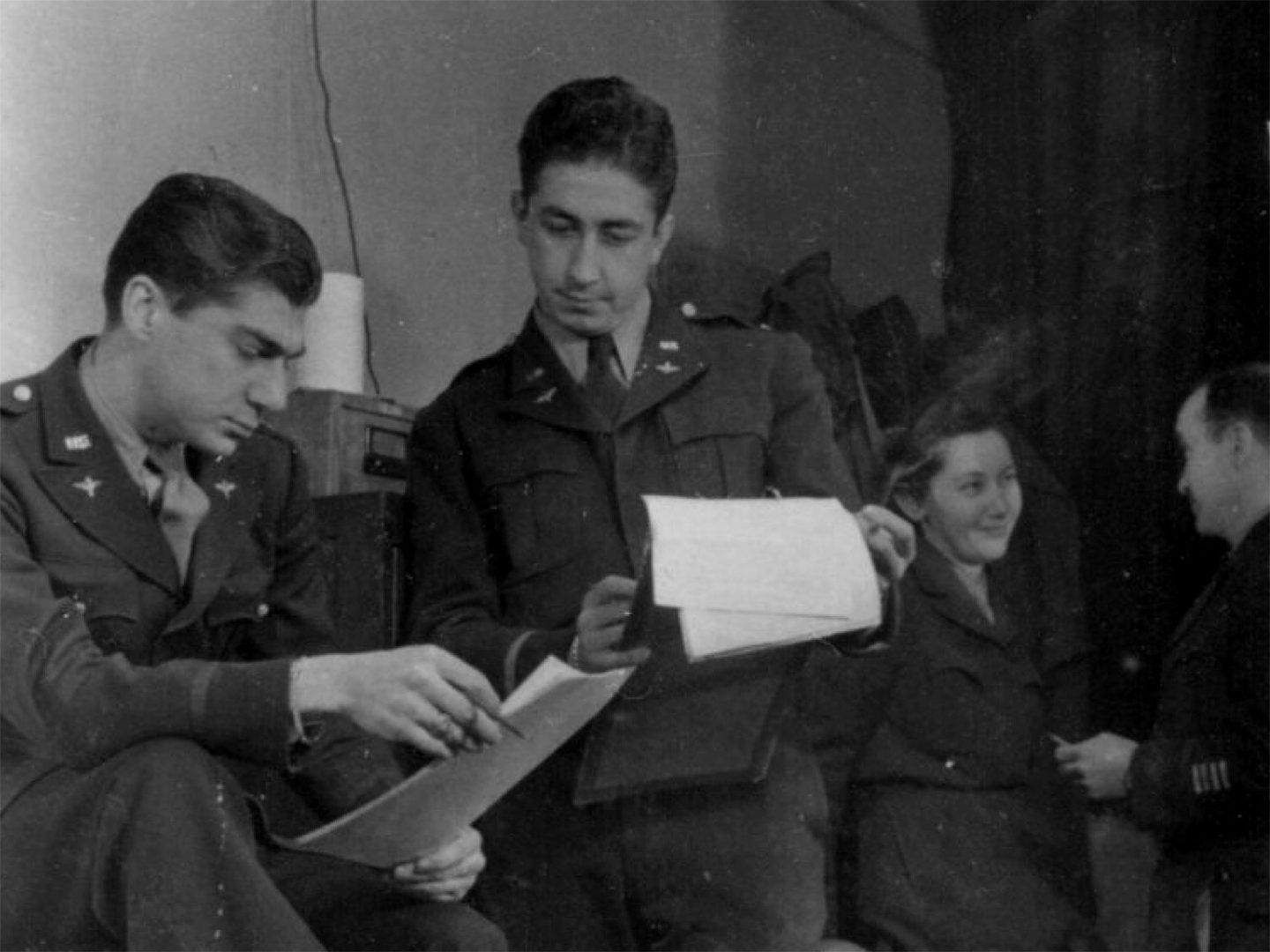 A historic black and white photo of two men with dark hair in US Air Force uniforms consulting some paperwork. One man holds a smoking cigarette. In the background a third man and a woman are talking.