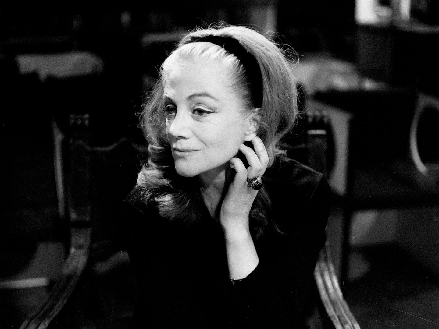 A black and white historic photo of a middle-aged woman seated in a chair. Her hair is pulled back by a black head band her left hand is raised to her face.