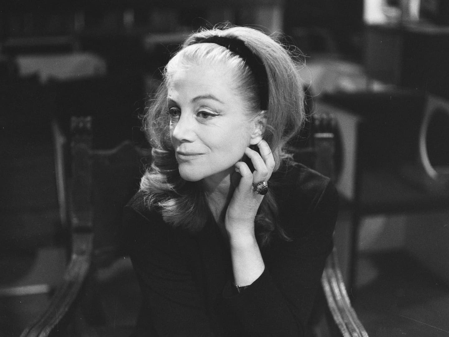A black and white historic photo of a middle-aged woman seated in a chair. Her hair is pulled back by a black head band her left hand is raised to her face.