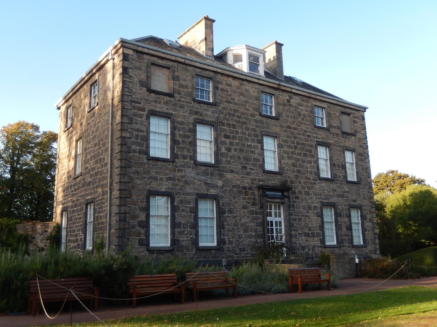 A modern colour photo of a Georgian-style house. It has 3 floors and a central doorway. Windows are arranged symmetrically either side of and above the doorway. Benches and a lawn are visible in front of the house.