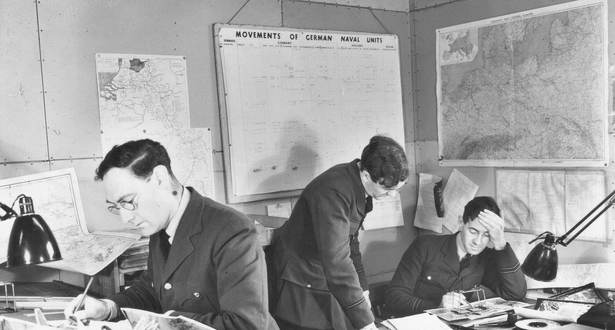 A historic black and white photo of 3 men in uniform in an office. To the left a man in glasses leans over a desk holding a pencil. To the right, 2 other men study a photo under a pocket stereoscope. On the walls behind them are maps and other paperwork relating to shipping.