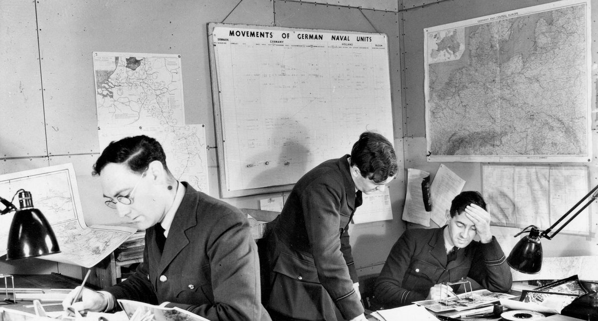 A historic black and white photo of 3 men in uniform in an office. To the left a man in glasses leans over a desk holding a pencil. To the right, 2 other men study a photo under a pocket stereoscope. On the walls behind them are maps and other paperwork relating to shipping.