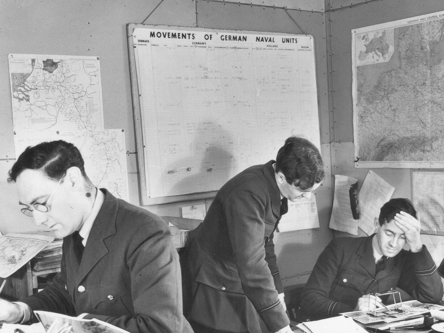 A historic black and white photo of 3 men in uniform in an office. To the left a man in glasses leans over a desk holding a pencil. To the right, 2 other men study a photo under a pocket stereoscope. On the walls behind them are maps and other paperwork relating to shipping.