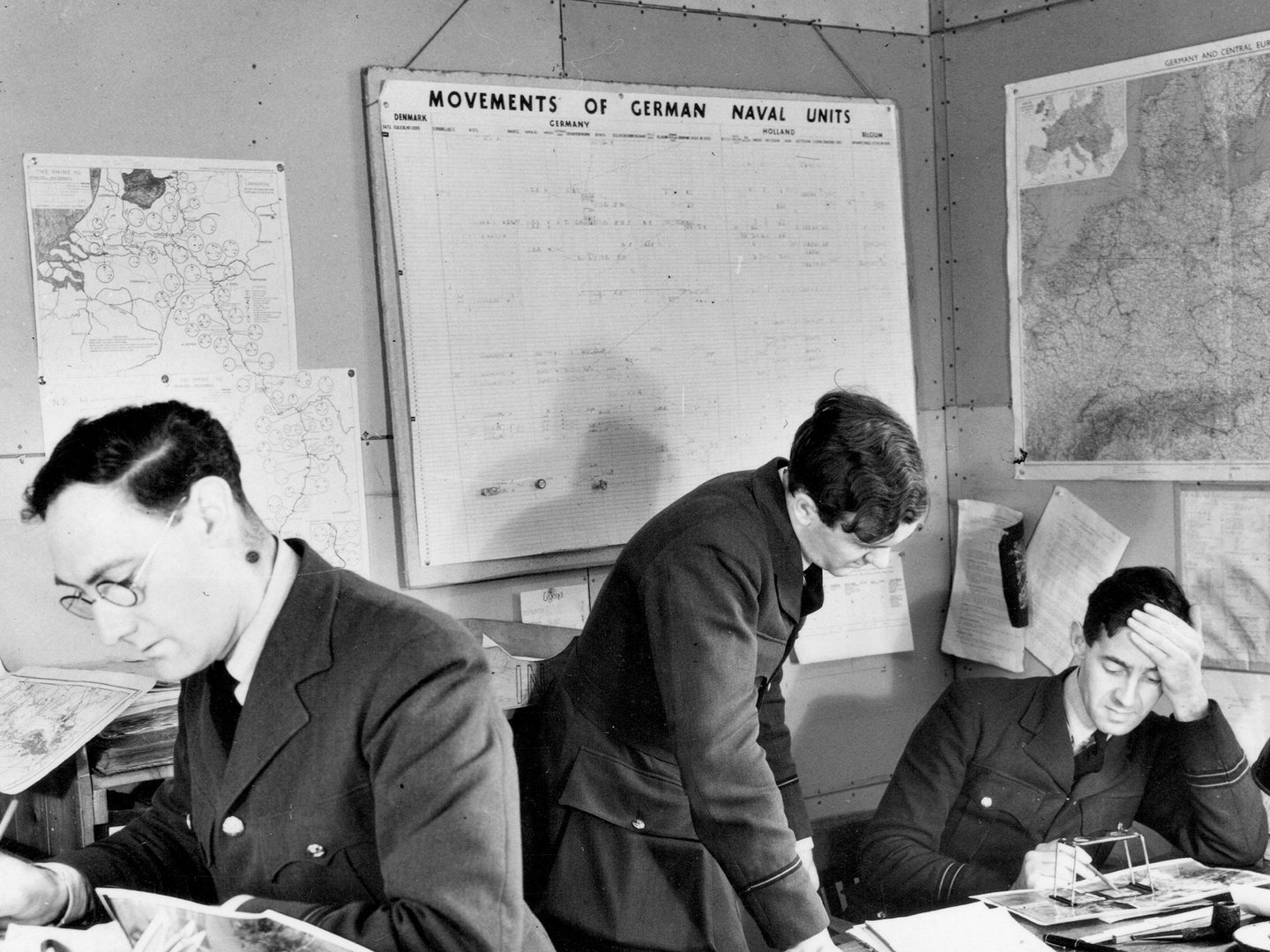 A historic black and white photo of 3 men in uniform in an office. To the left a man in glasses leans over a desk holding a pencil. To the right, 2 other men study a photo under a pocket stereoscope. On the walls behind them are maps and other paperwork relating to shipping.