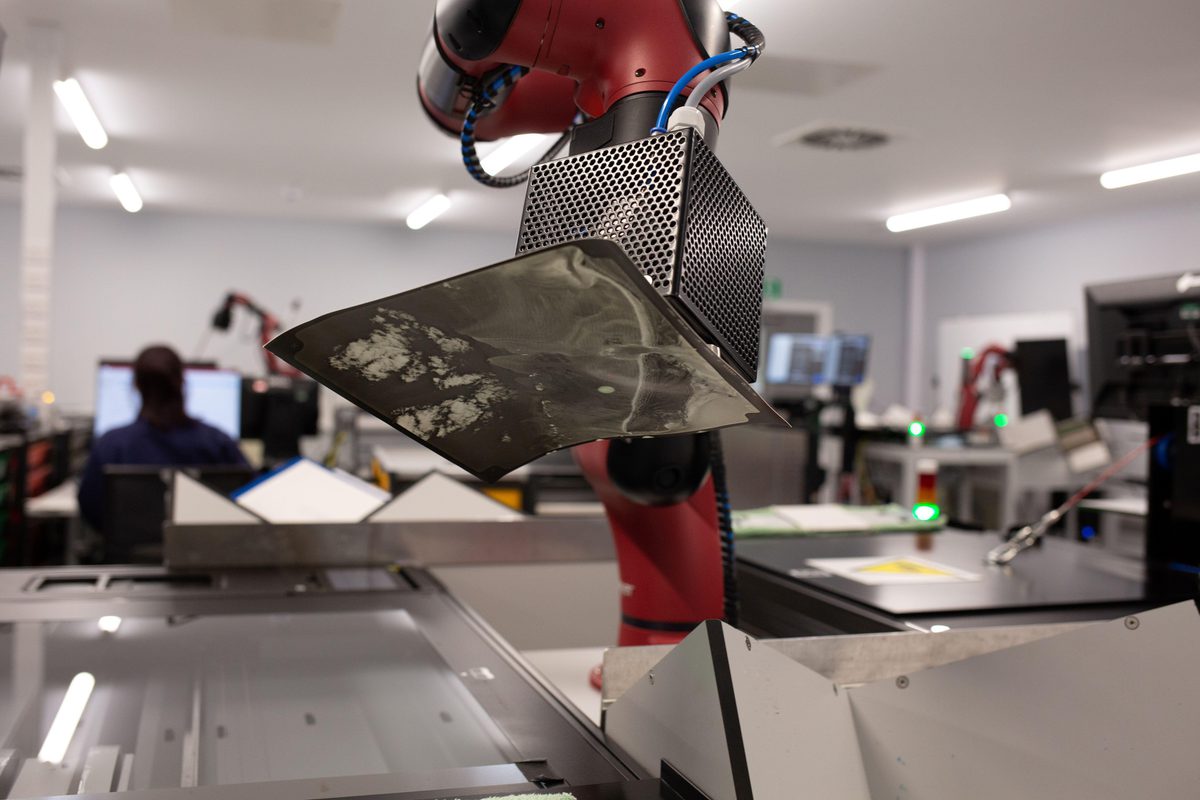 A red robot arm carefully lifts a historic photographic print onto a glass scanner bed. The background is out of focus, but other robot arms and scanners are just visible. To the left, a person sits at a computer screen with their back to the camera.