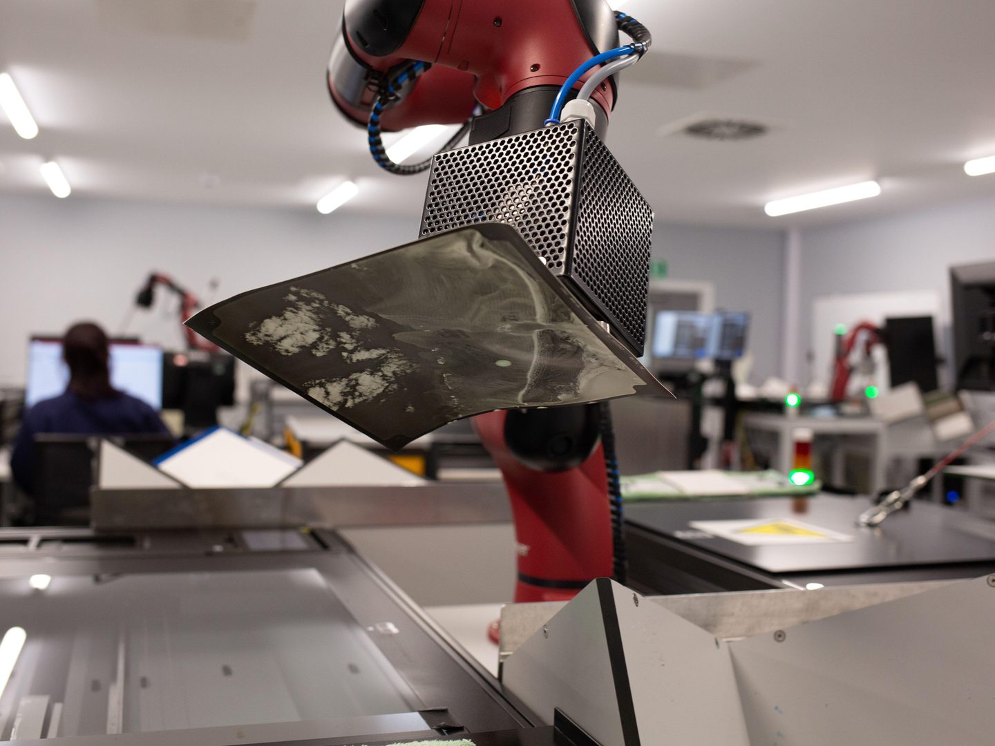 A red robot arm carefully lifts a historic photographic print onto a glass scanner bed. The background is out of focus, but other robot arms and scanners are just visible. To the left, a person sits at a computer screen with their back to the camera.
