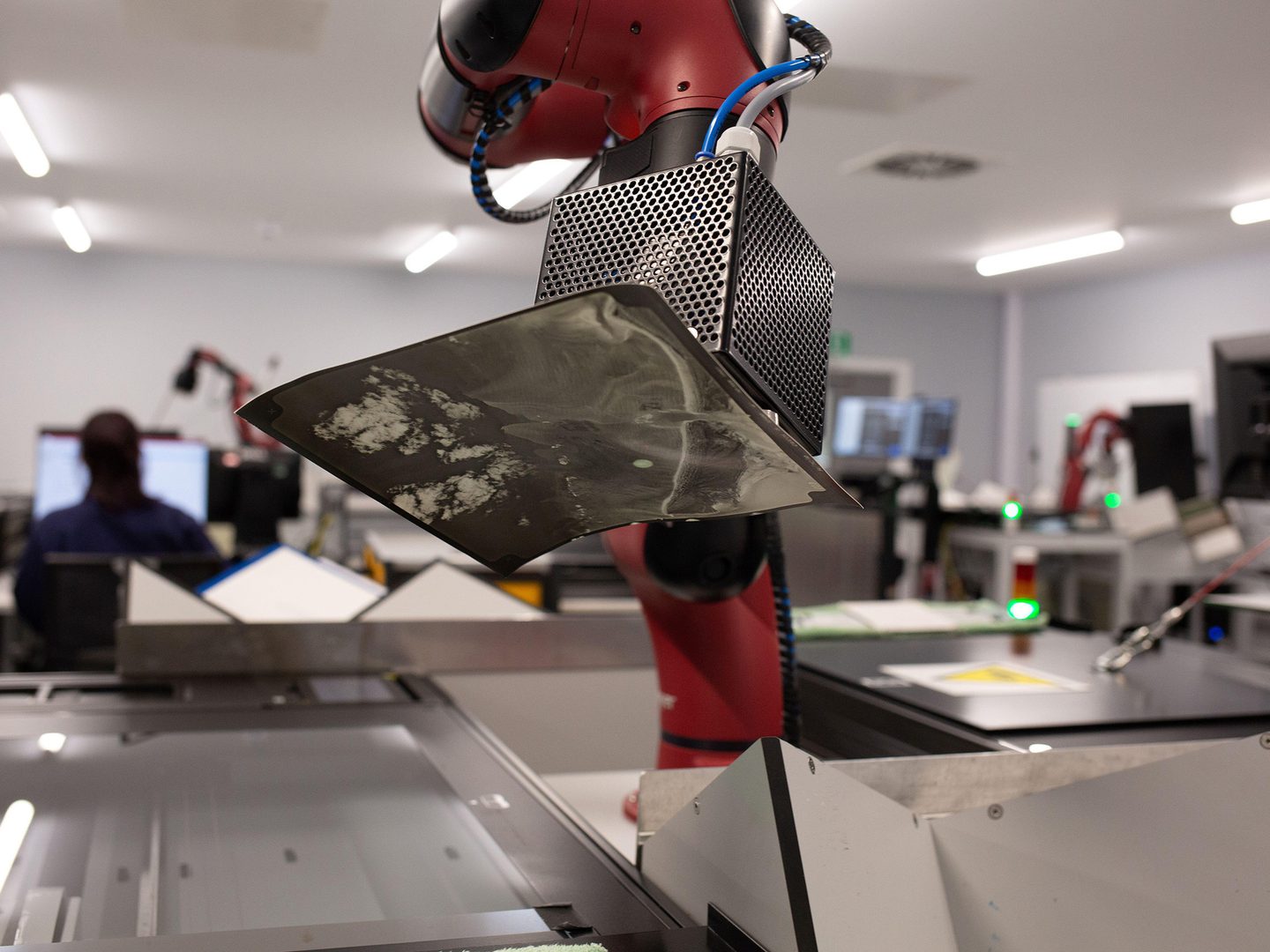 A red robot arm carefully lifts a historic photographic print onto a glass scanner bed. The background is out of focus, but other robot arms and scanners are just visible. To the left, a person sits at a computer screen with their back to the camera.