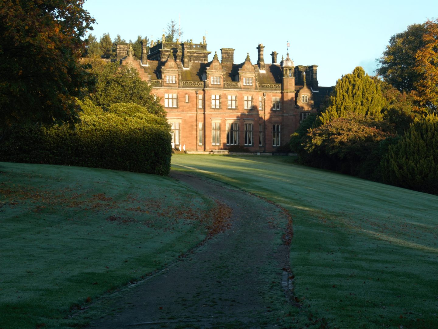 A grand red brick building with cupulas at each corner. A driveway leads from the photographer towards the building, flanked by frost-covered lawns scattered with fallen leaves. Trees are visible either side of the driveway, and behind the building.