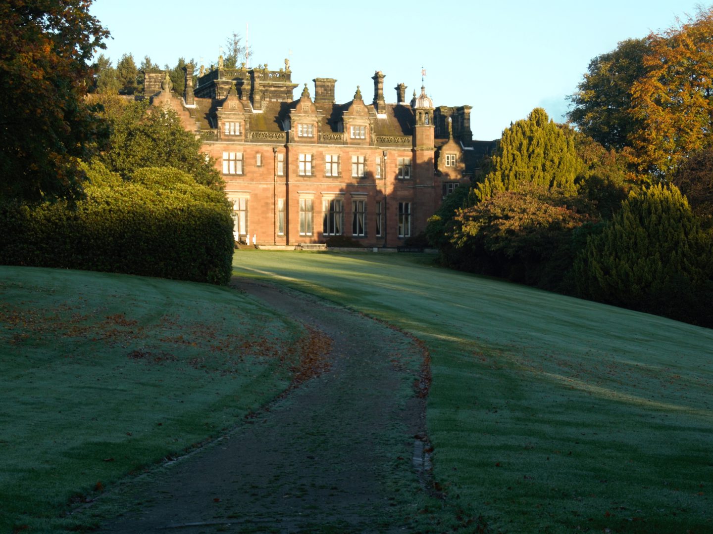 A grand red brick building with cupulas at each corner. A driveway leads from the photographer towards the building, flanked by frost-covered lawns scattered with fallen leaves. Trees are visible either side of the driveway, and behind the building.
