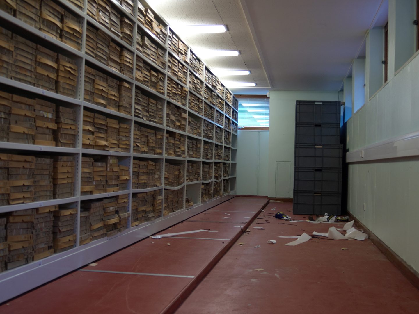 A strip-lit room with white walls and a red floor. Floor-to-ceiling metal shelving occupies the left side, stuffed with hundreds of slim cardboard boxes stacked on top of each other. At the far end of the room is a stack of large grey plastic boxes.