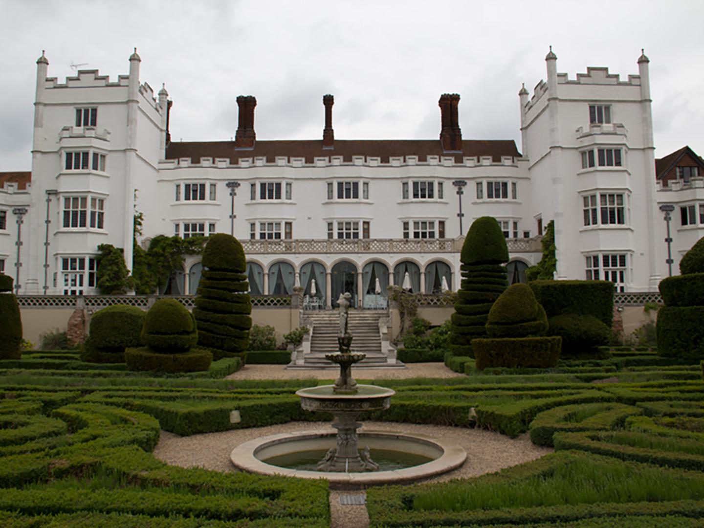 A grand white stately home with rows of regular windows and red tiled chimneys. Topiary bushes and a fountain occupy the foreground.