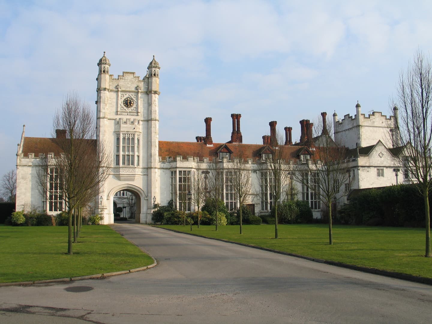 A grand white stately home with a tower, rows of regular windows and red tiled chimneys. A drive flanked by young trees leads up to the house.